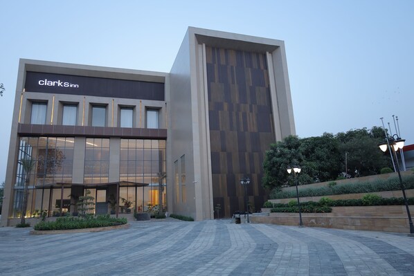 A modern Clark's Inn building with a large glass facade, a wooden panel design on the side, and a paved courtyard with plants and street lamps. - Clarks Inn Etawah (Etawah)
