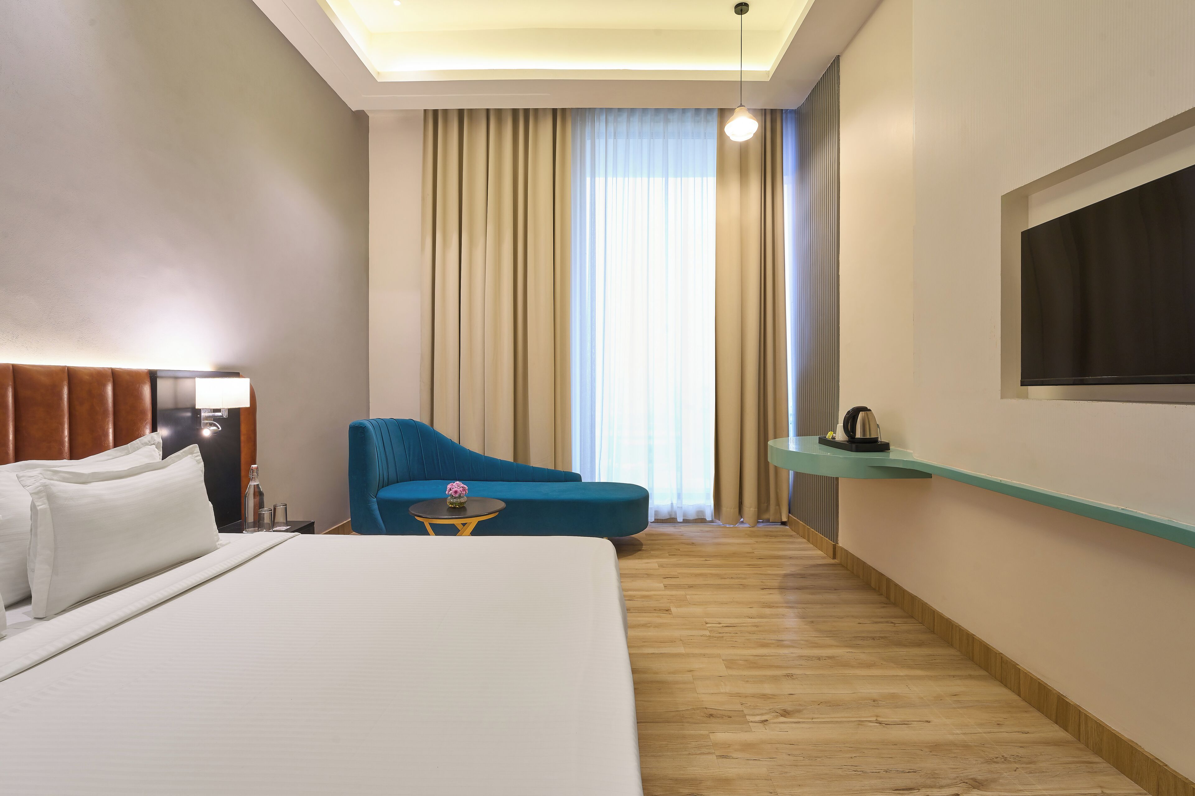 Una habitación de hotel con una cama grande, un sillón azul, una mesita de noche con una lámpara y un televisor fijado a la pared.