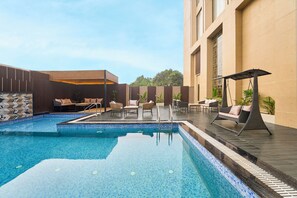 A swimming pool with lounge chairs and a sunbed, surrounded by a building with large windows and a tiled floor. - Clarks Inn Etawah (Etawah)