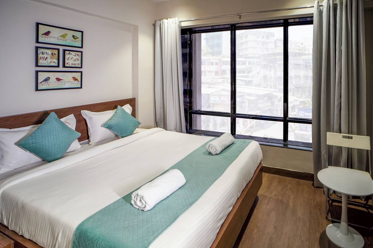 A neatly made bed with a green and white bedspread, a white pillow, and a rolled white towel. There is a small white table and a chair to the right. The room has a large window with curtains, and a wall with framed pictures of birds.