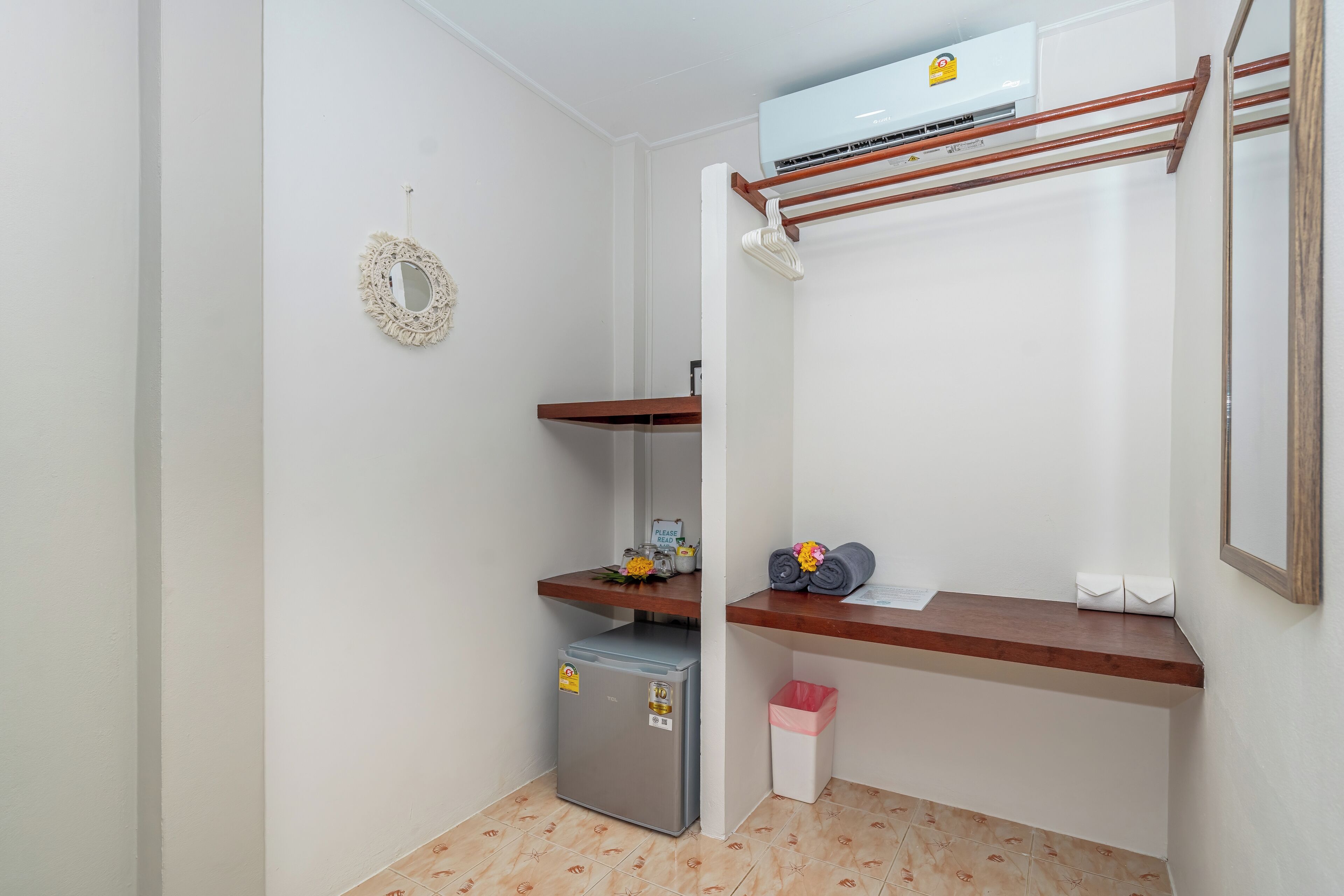 Et lille værelse med indbygget klimaanlæg, et minikøleskab, en træhylde med toiletartikler, en skraldespand og et spejl på væggen.