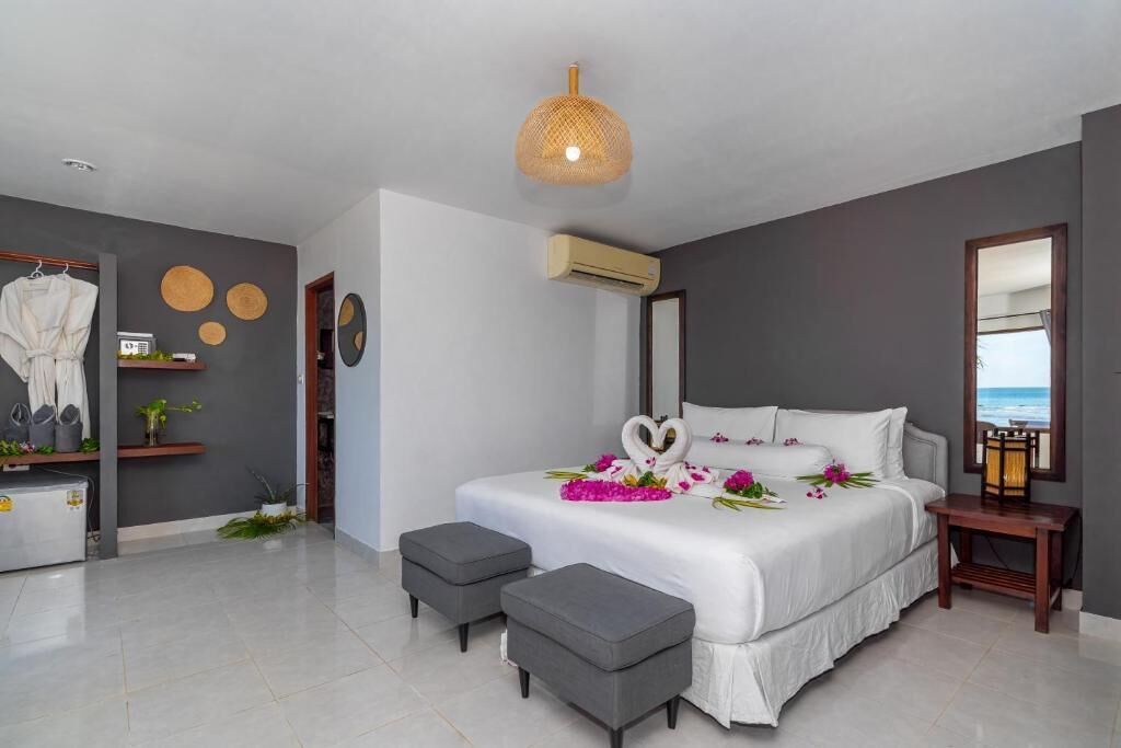 A modern bedroom with a large bed, grey ottomans, a wall-mounted air conditioner, and a beach view through a window.