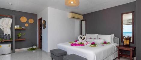 A modern bedroom with a large bed, grey ottomans, a wall-mounted air conditioner, and a beach view through a window.