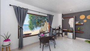Un soggiorno moderno con una grande finestra che si affaccia su una spiaggia, una zona pranzo con sedie in vimini e un angolo cottura con frigorifero e scaffali.