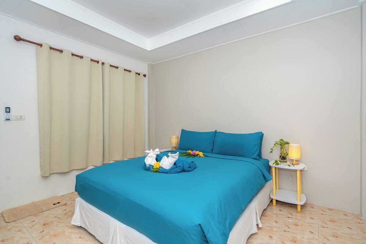 A neatly made bed with blue bedding, a bedside table with a lamp, and a window with curtains.