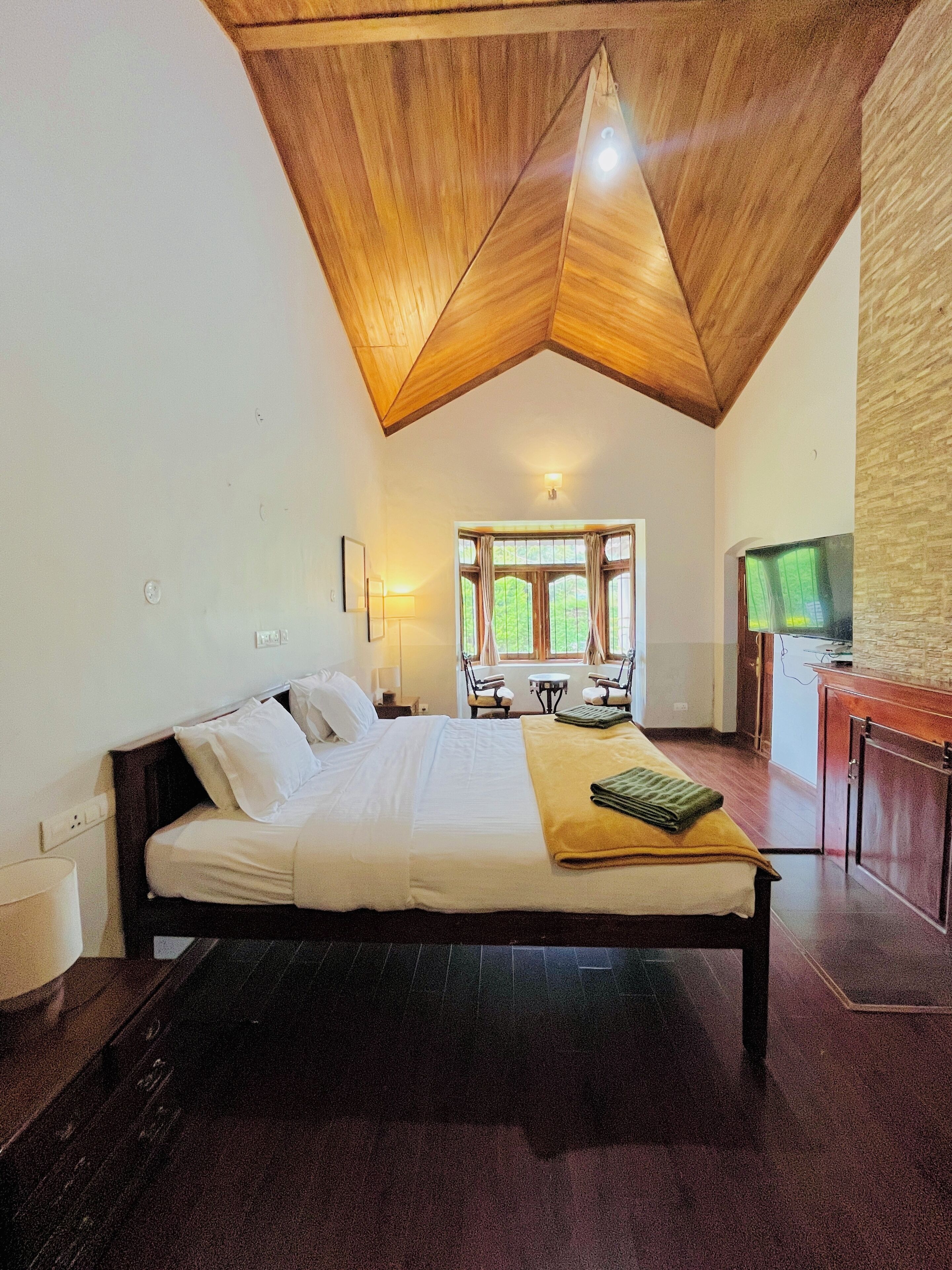 A hotel room with a wooden ceiling, a bed, a desk, a TV, and a window with curtains.