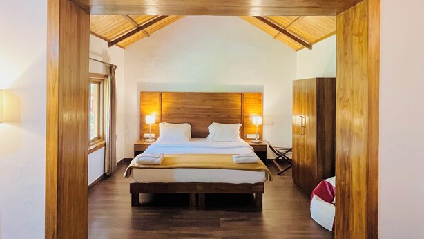 A bedroom with a wooden ceiling, a large bed, bedside lamps, and a view through an open doorway. - BouganVilla (Ootacamund)
