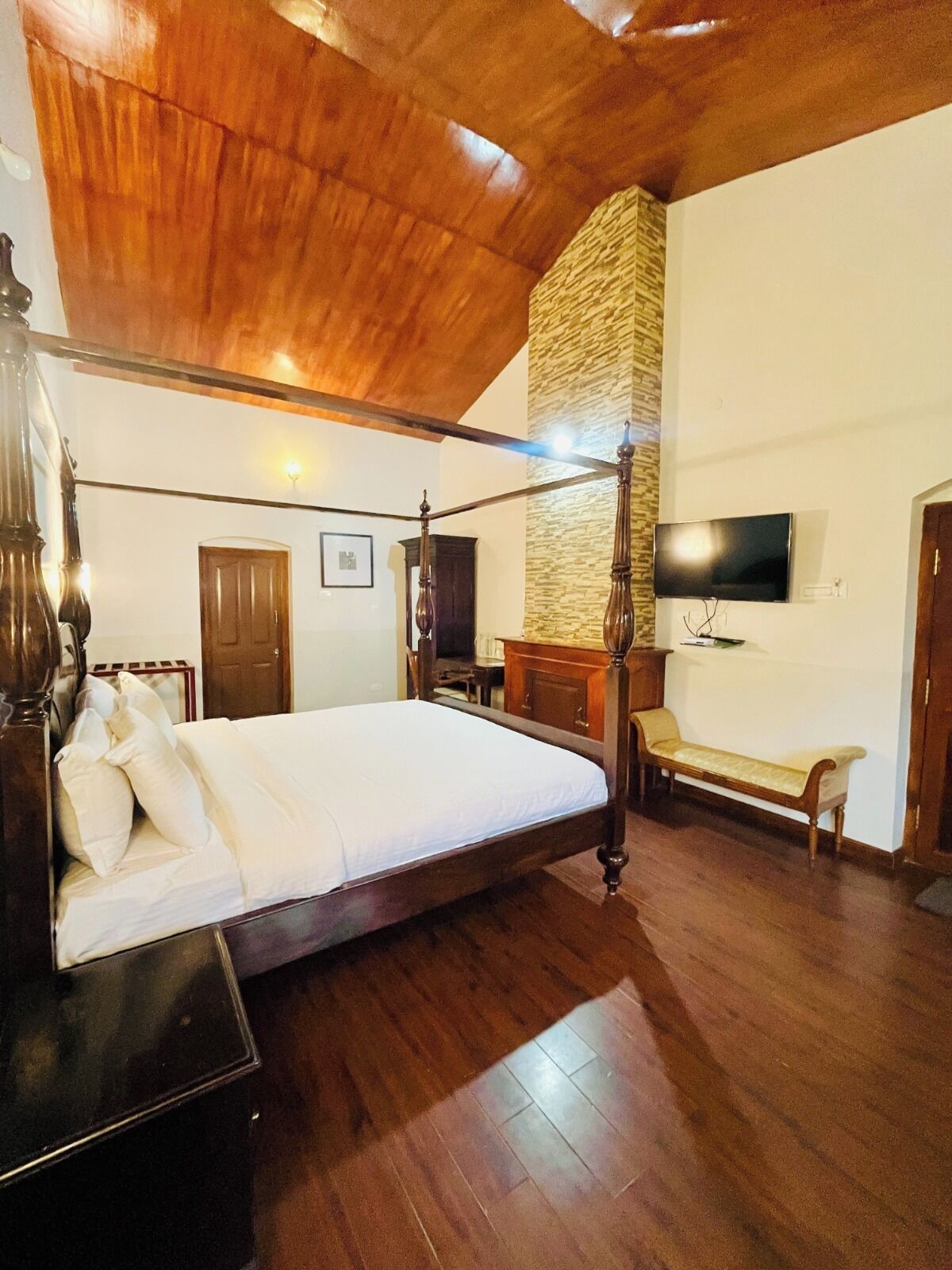 Un dormitorio amplio con una cama grande, techo de madera, pared de acento de piedra, televisor y una banca.