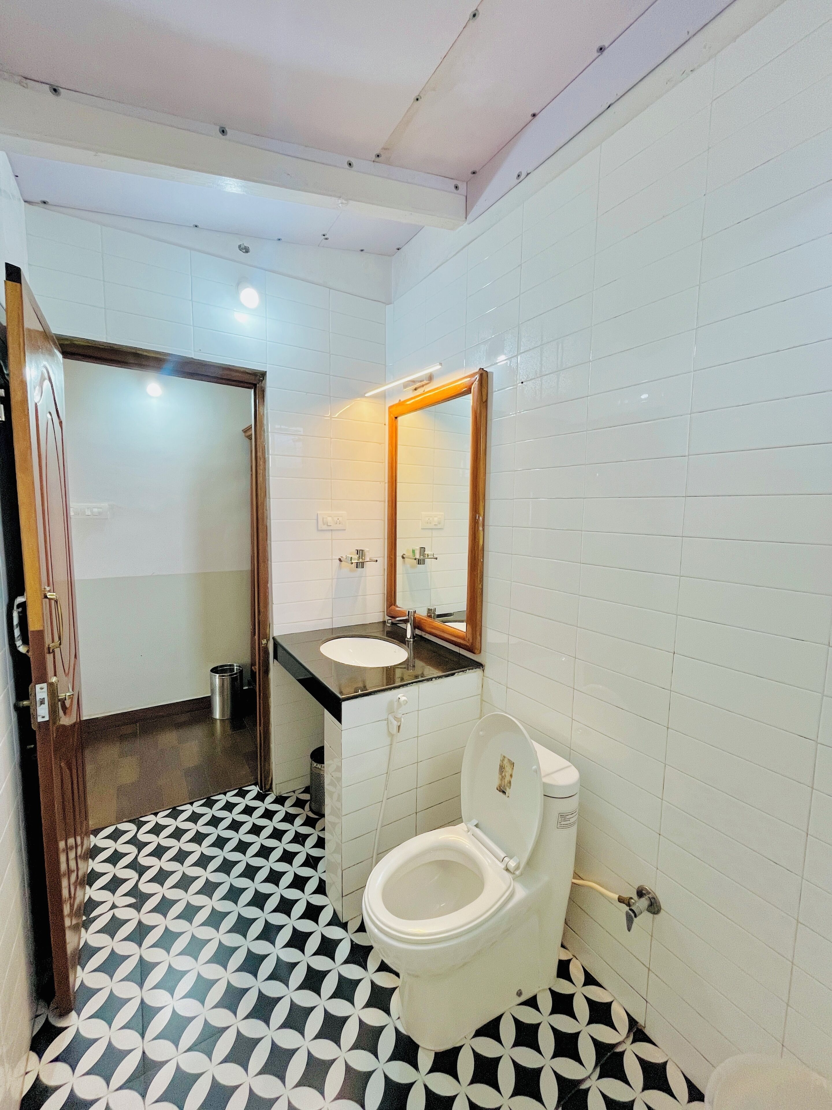 A bathroom with white tiled walls, a black and white patterned floor, a white toilet, a sink with a wooden frame mirror, and a door leading to another room.