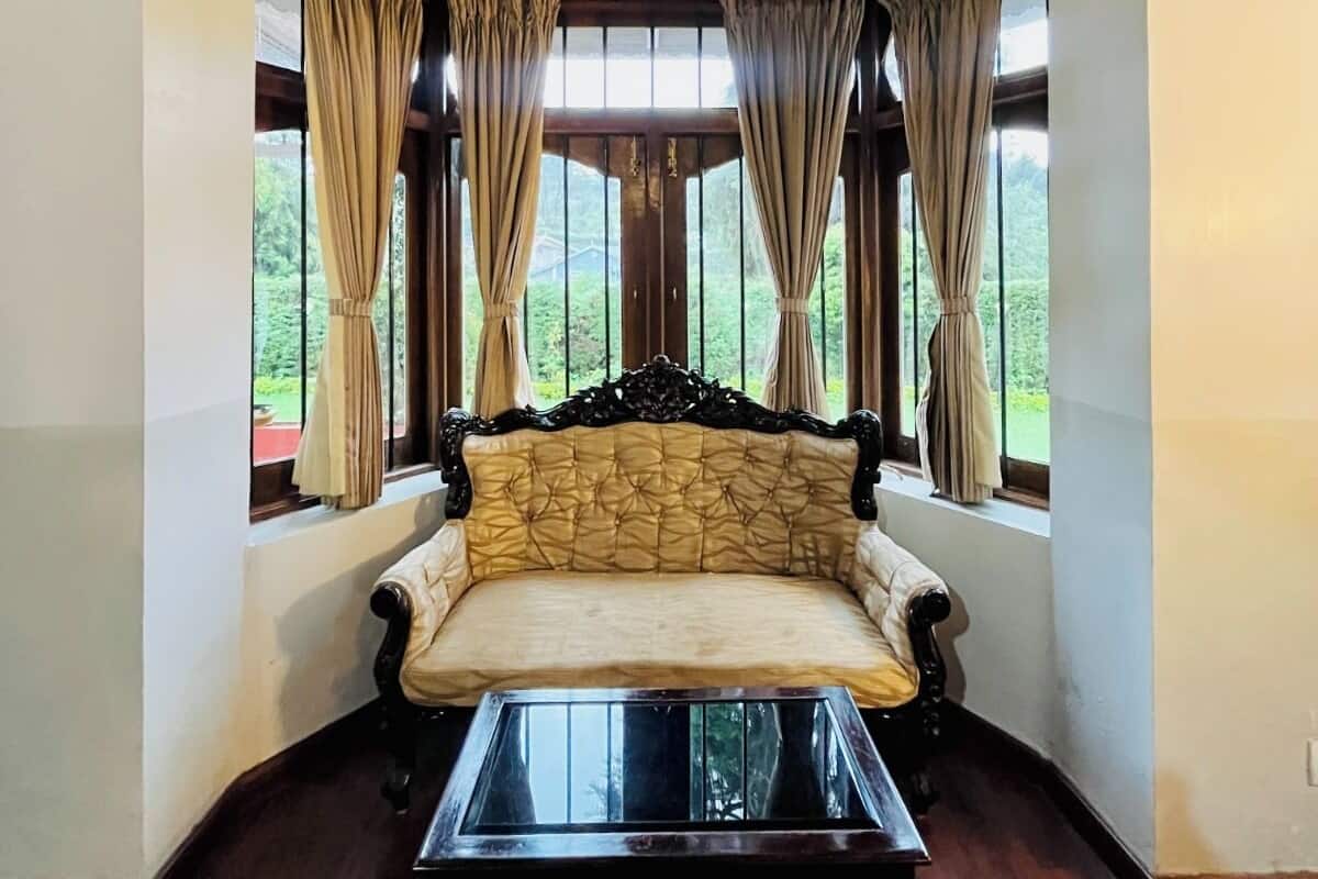 A corner room with a sofa and a glass coffee table, large windows with curtains, and wooden flooring.