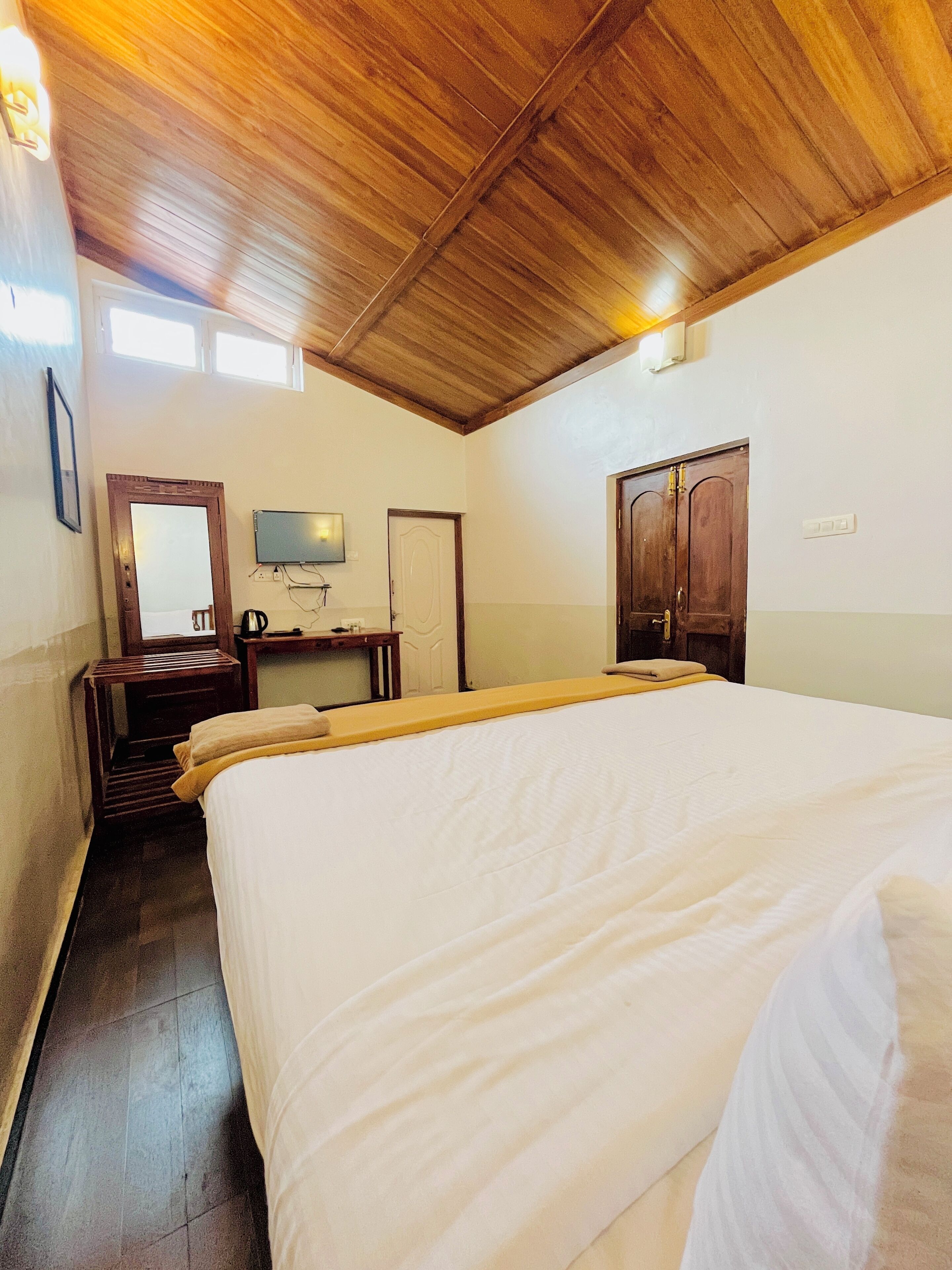 Un dormitorio con techo de madera, una cama con ropa de cama blanca, un escritorio con silla, un televisor y un armario.