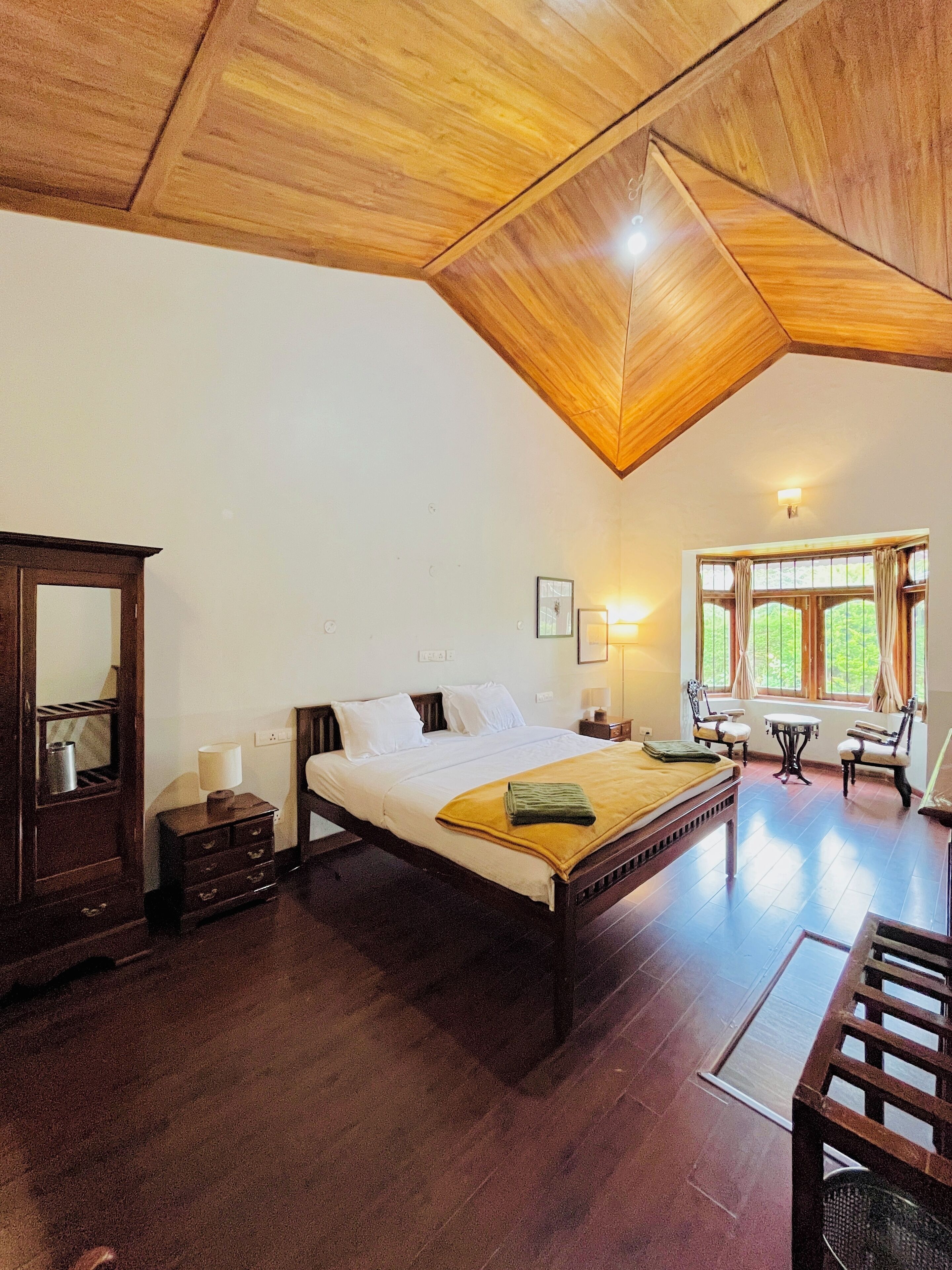 A room with a wooden ceiling, a bed with white linens, a nightstand, a dresser, a window with curtains, and a small table with chairs.