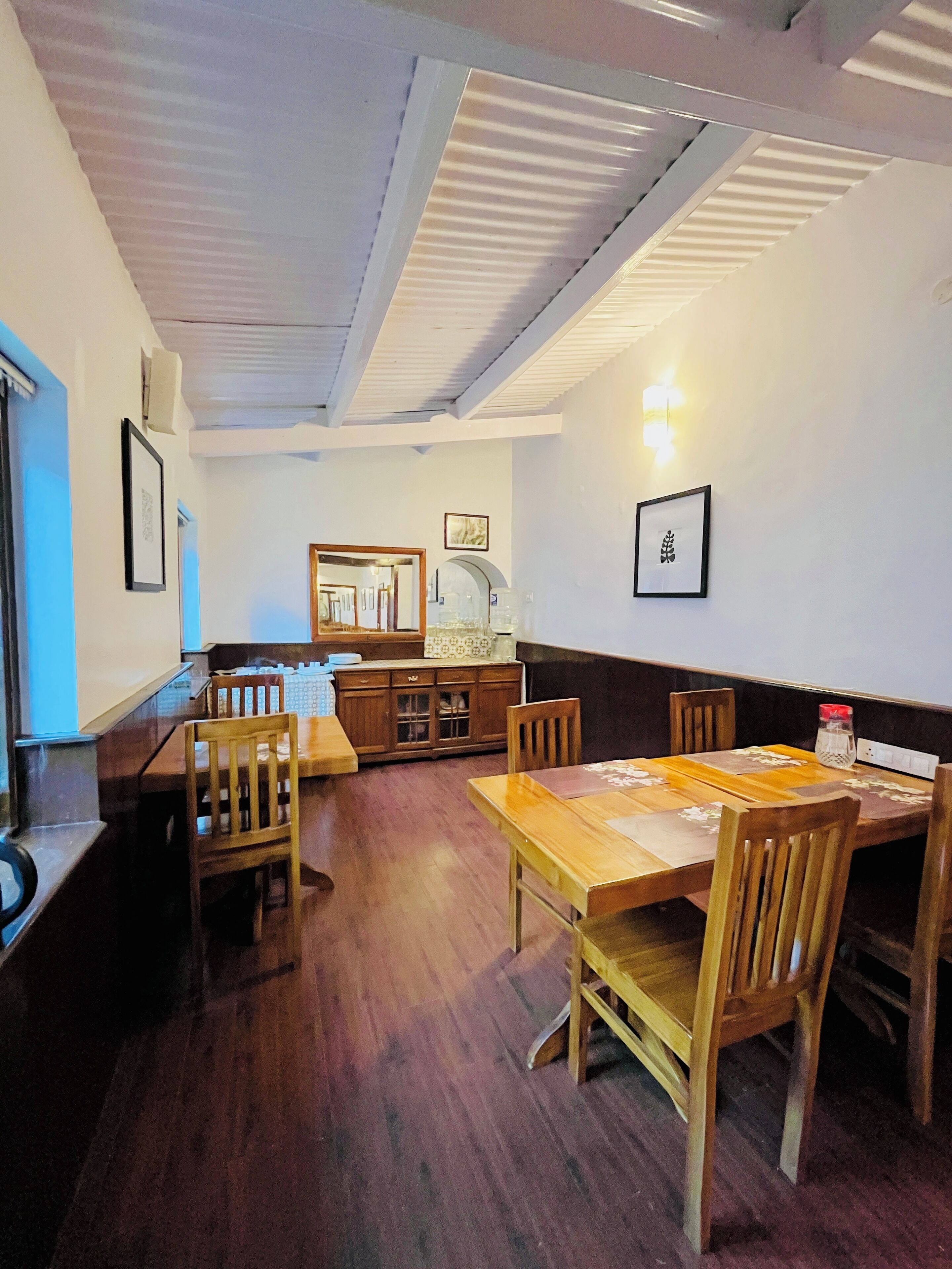 Un espace repas avec des tables et des chaises en bois, une cuisine à l’arrière-plan et des tableaux encadrés accrochés aux murs.