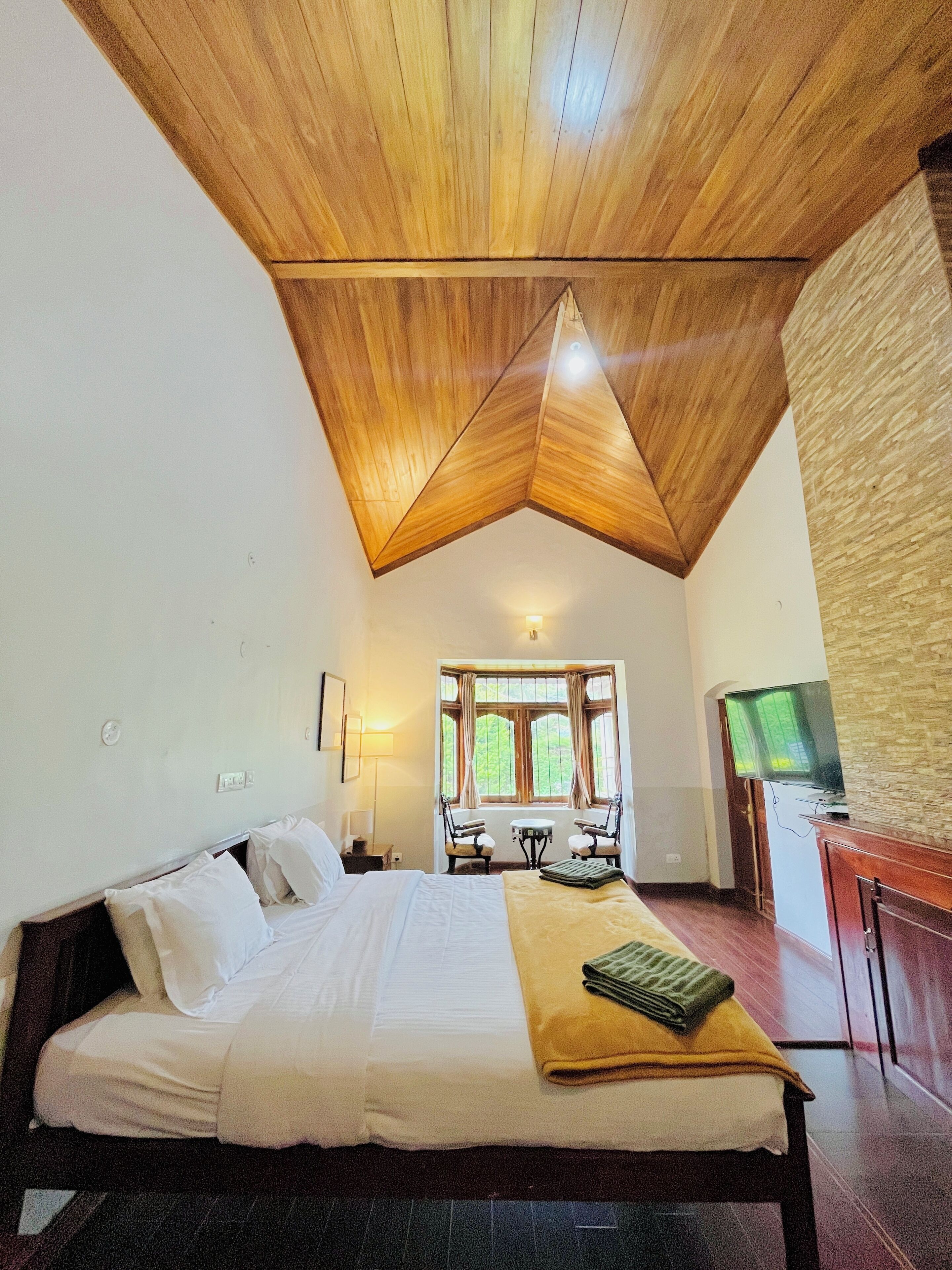 Un dormitorio con techo de madera, una cama con ropa de cama blanca, una mesa con sillas, un televisor y un ventanal con cortinas.