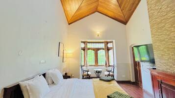 Un dormitorio con techo de madera, una cama con ropa de cama blanca, una mesa con sillas, un televisor y un ventanal con cortinas.