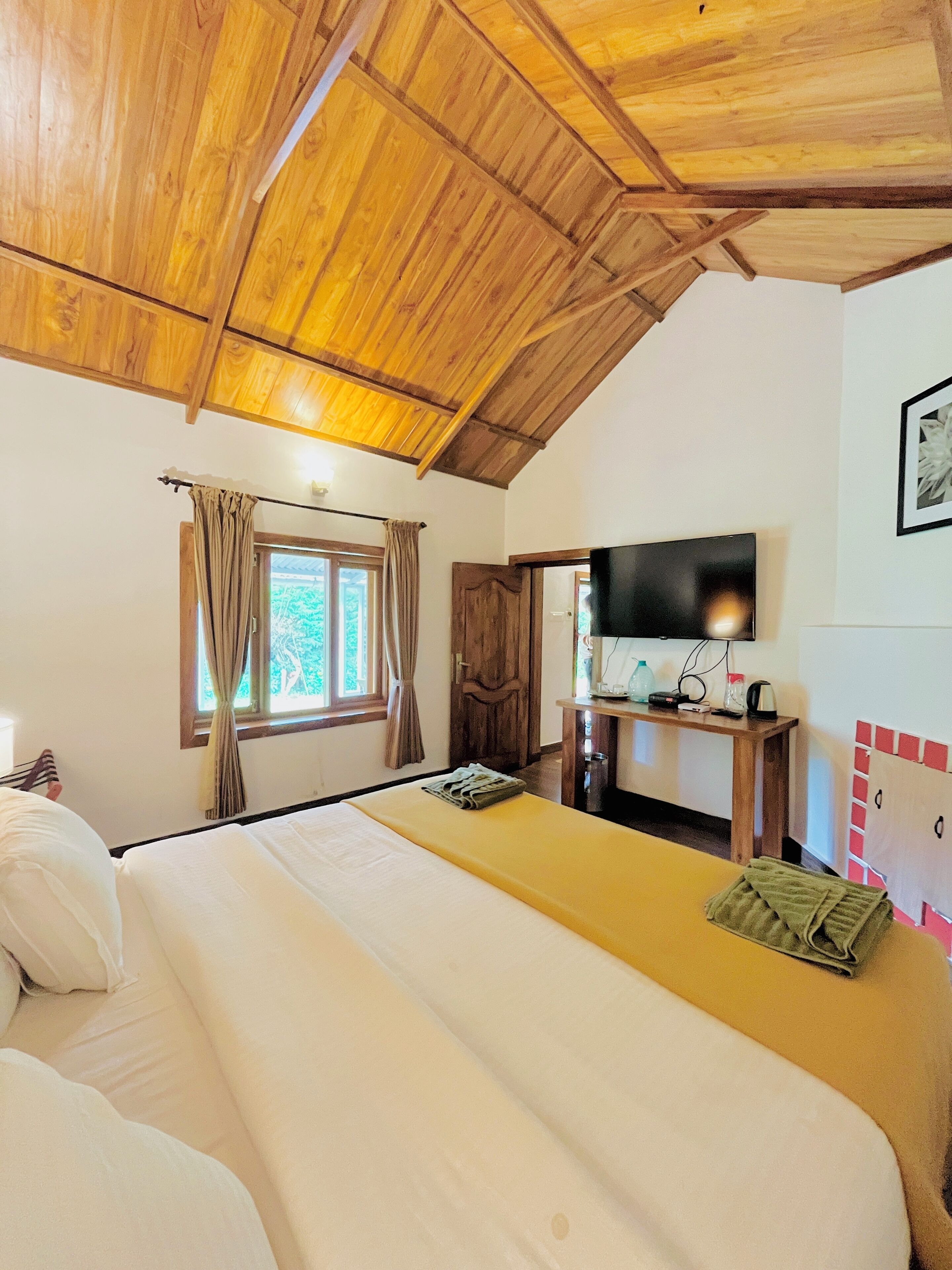 Un dormitorio con techo de madera, una cama con ropa de cama blanca y amarilla, un televisor sobre un mueble de madera y una ventana con cortinas.