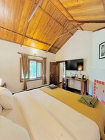 Un dormitorio con techo de madera, una cama con ropa de cama blanca y amarilla, un televisor sobre un mueble de madera y una ventana con cortinas.