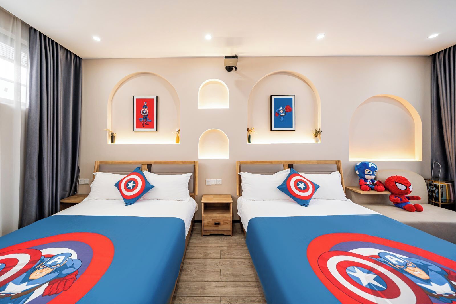 Habitación de hotel con dos camas, cada una con una colcha decorada con un diseño de Capitán América. Hay luces fijadas en la pared y carteles enmarcados sobre las camas.