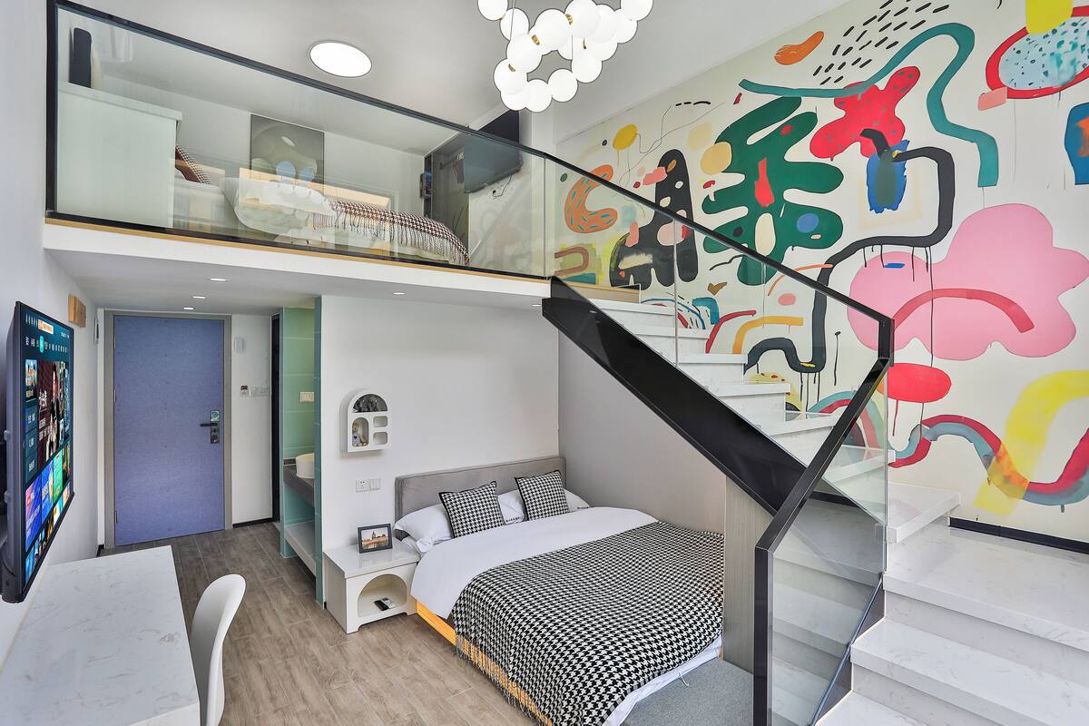 A modern bedroom with a bed, a desk, a chair, a staircase, and a colorful abstract wall mural.