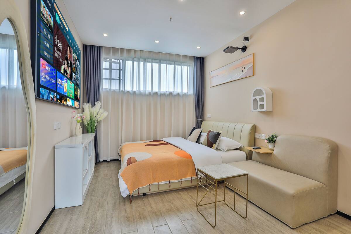 A modern hotel room with a bed, a sofa, a desk, and a TV displaying various channels.