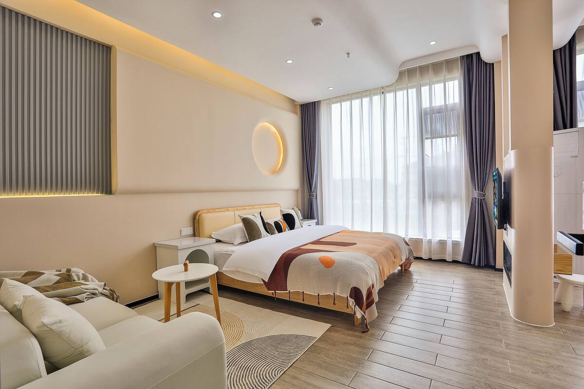 A modern hotel room with a large bed, a sitting area with a sofa and armchair, a small table, and a TV.