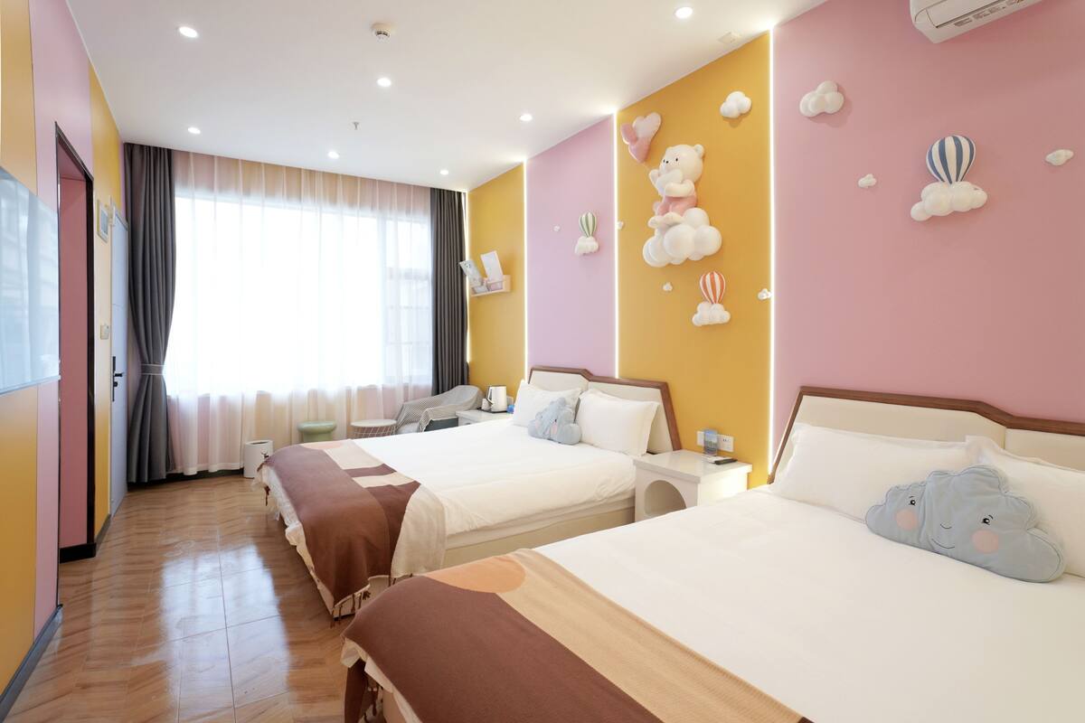 A hotel room with two beds, a large window with curtains, a bedside table with a lamp, and a wall decorated with cloud and hot air balloon decals.