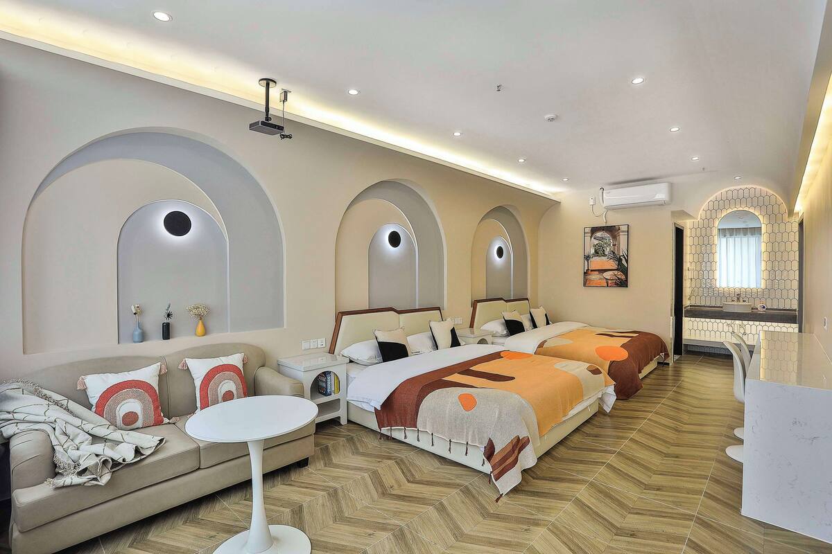 A hotel room with a sofa, two beds, a small table, and a bathroom with a mirror and sink.