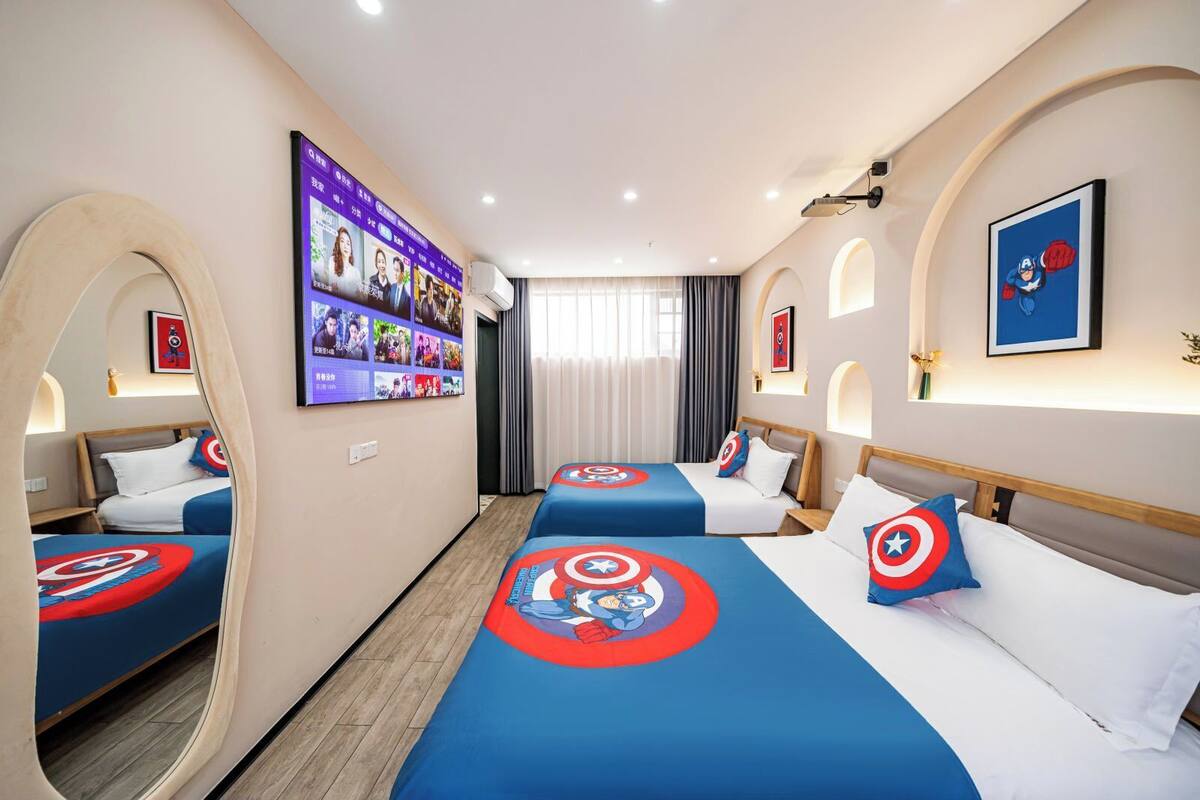 A hotel room with two beds, each with Captain America themed bedding, a large digital screen displaying various content, and a view of the outside through a window with curtains.