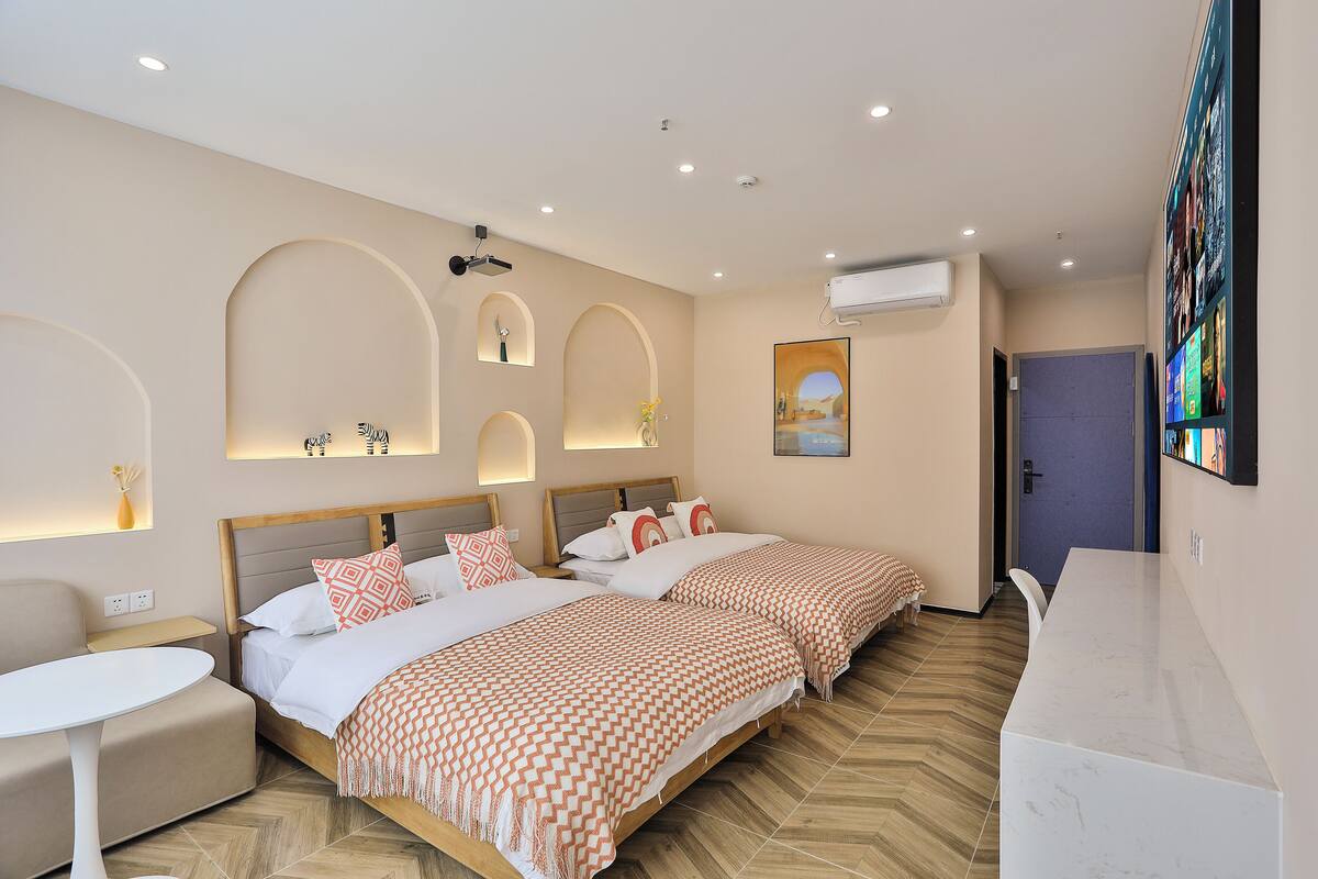 A modern hotel room with two beds, a round dining table, a built-in TV, and a large window.