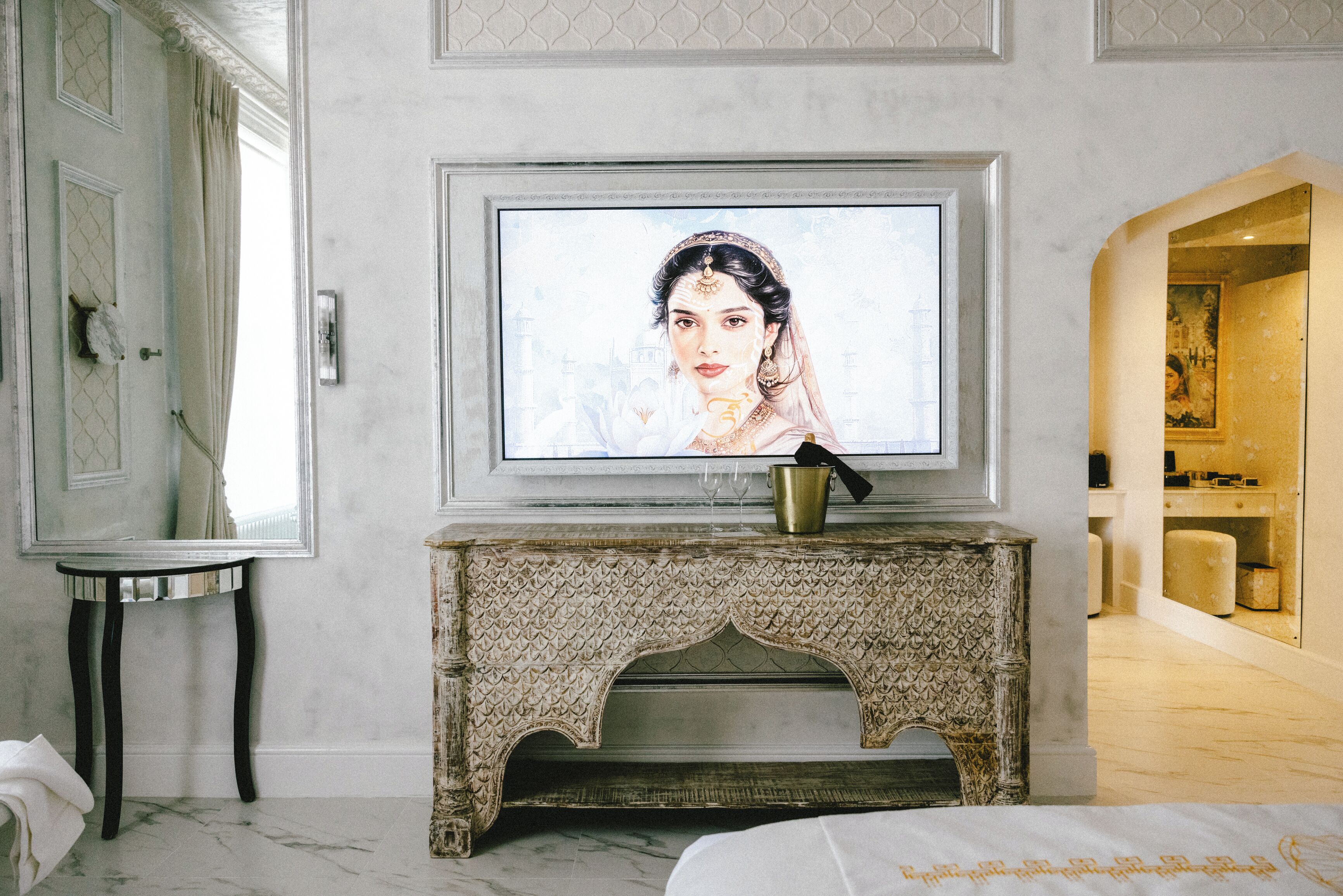 A room with a marble floor, a television mounted on a decorative fireplace, a small table with a mirror, and a doorway leading to another room.