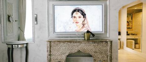 A room with a marble floor, a television mounted on a decorative fireplace, a small table with a mirror, and a doorway leading to another room.