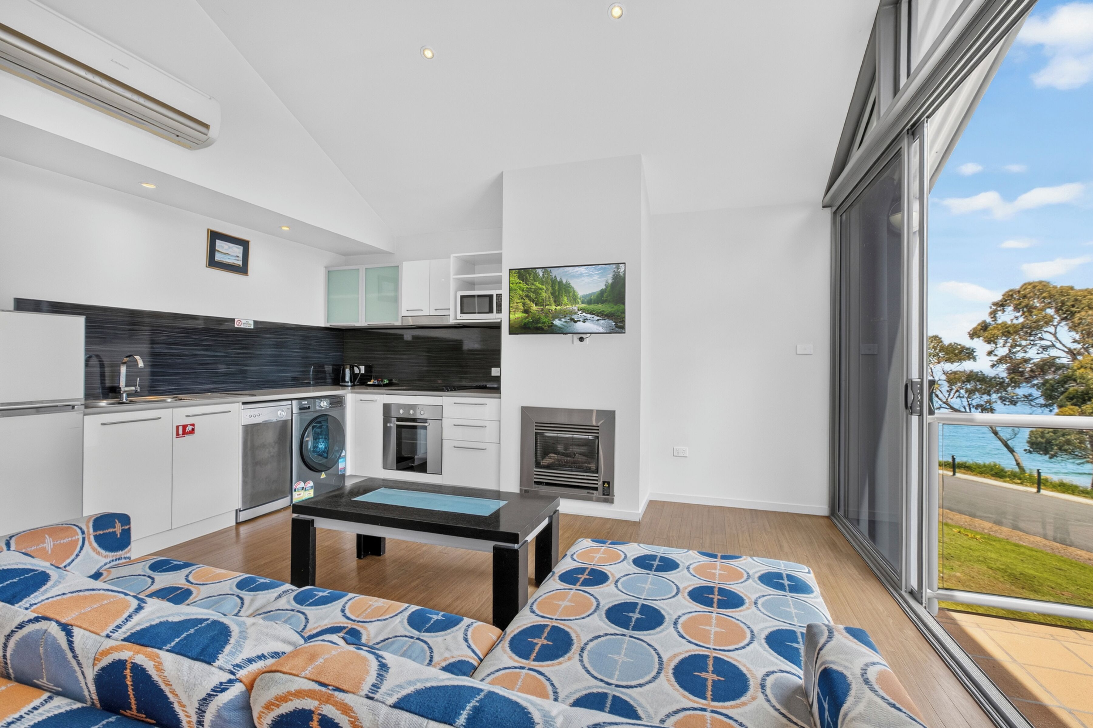 Una cocina moderna con electrodomésticos integrados, un comedor con mesa y sofá estampado, y un amplio ventanal corredero a un espacio exterior con vistas panorámicas.