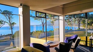 A modern room with large windows offering a view of the sea and trees. There is a round wooden table with two chairs and a small table with a chair. - Lufra Beachside Hotel (Eaglehawk Neck)