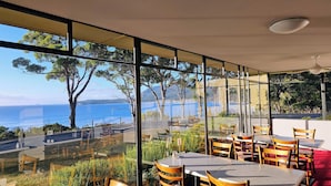 A restaurant with large windows offering a view of the sea and trees. Tables and chairs are arranged for dining. - Lufra Beachside Hotel (Eaglehawk Neck)