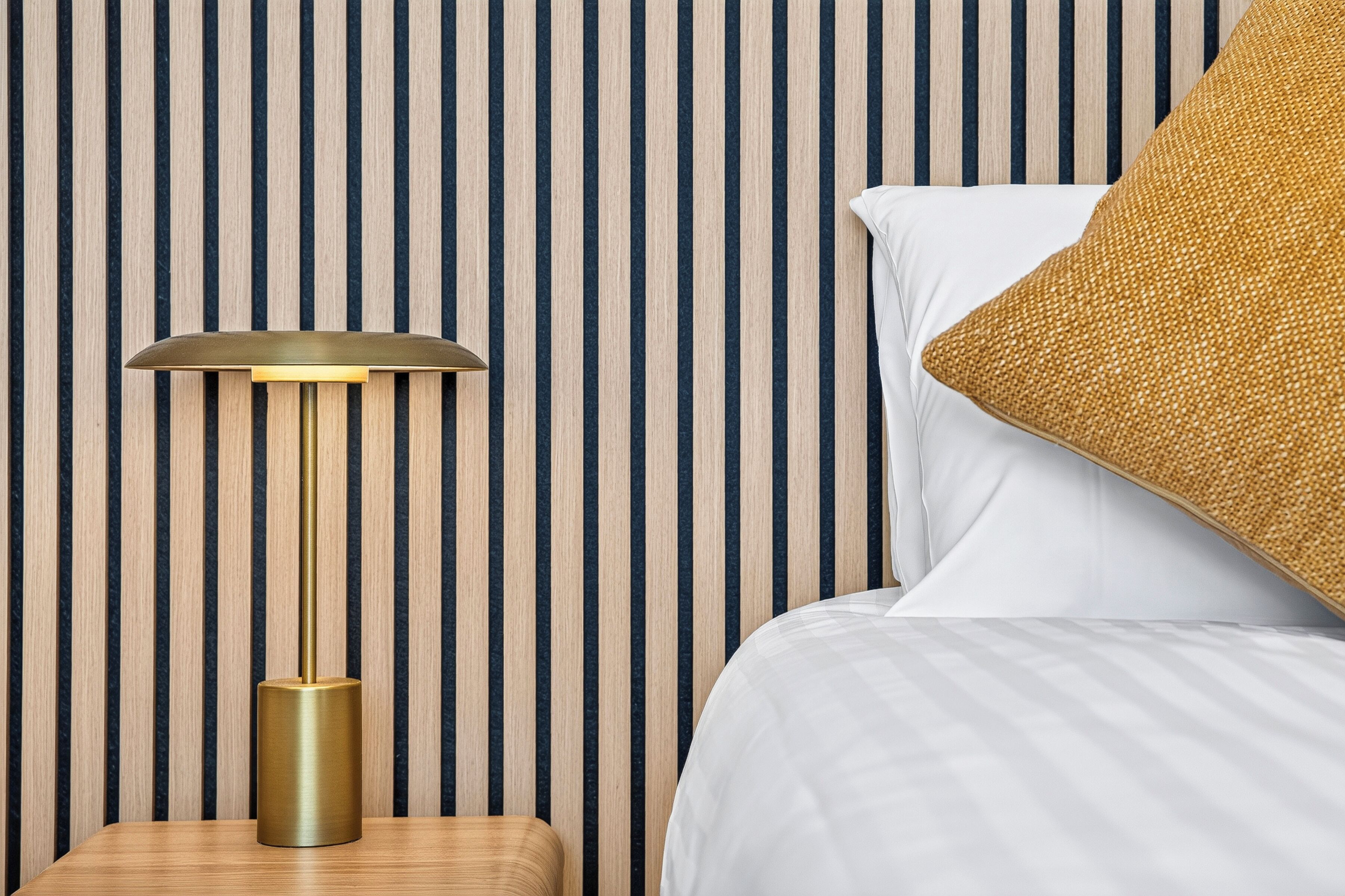 A bed with white linens, a gold-colored lamp on a wooden nightstand, and a striped wall.