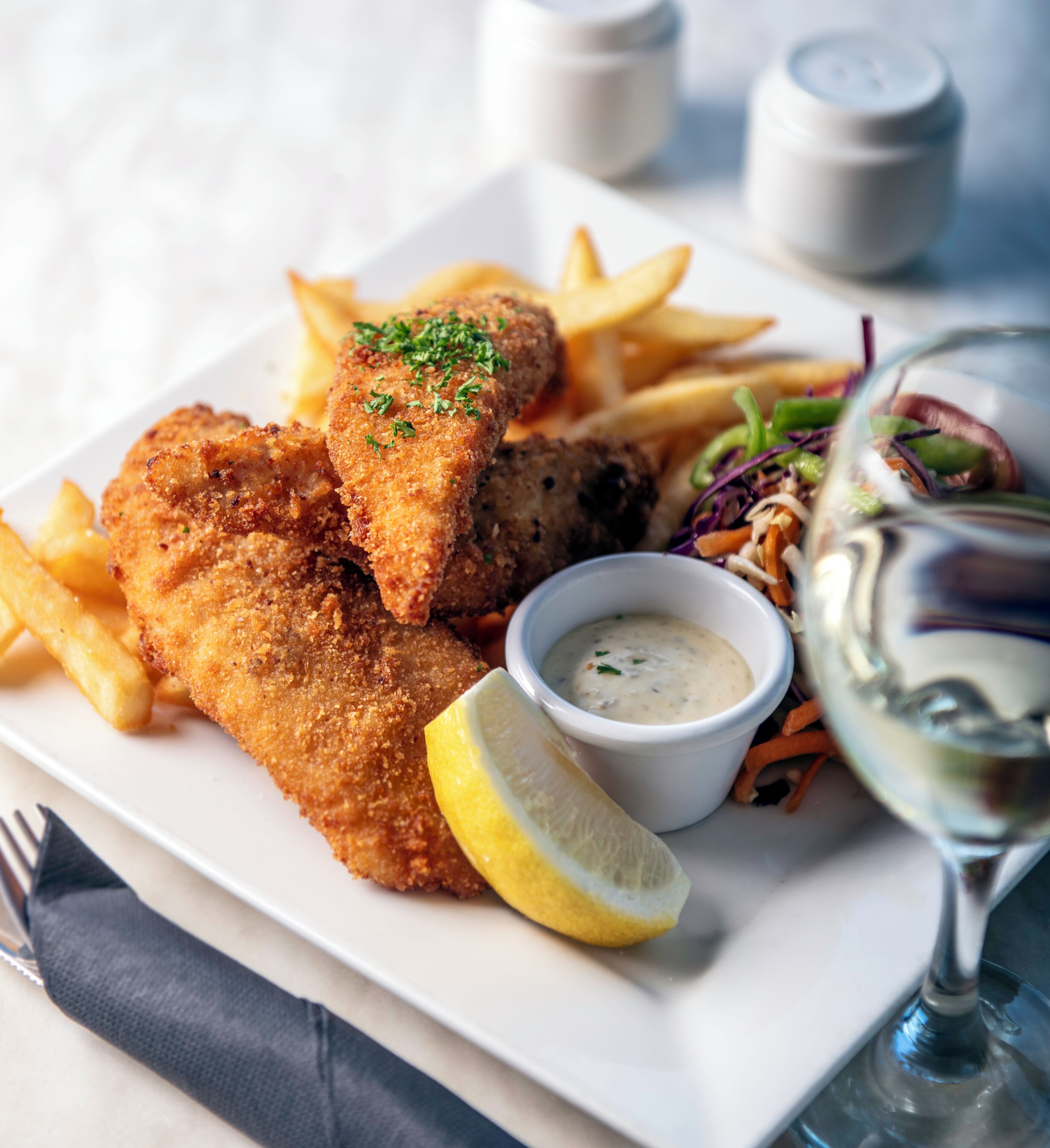A plate of breaded fish fillets, French fries, a lemon wedge, a small bowl of sauce, and a side salad.