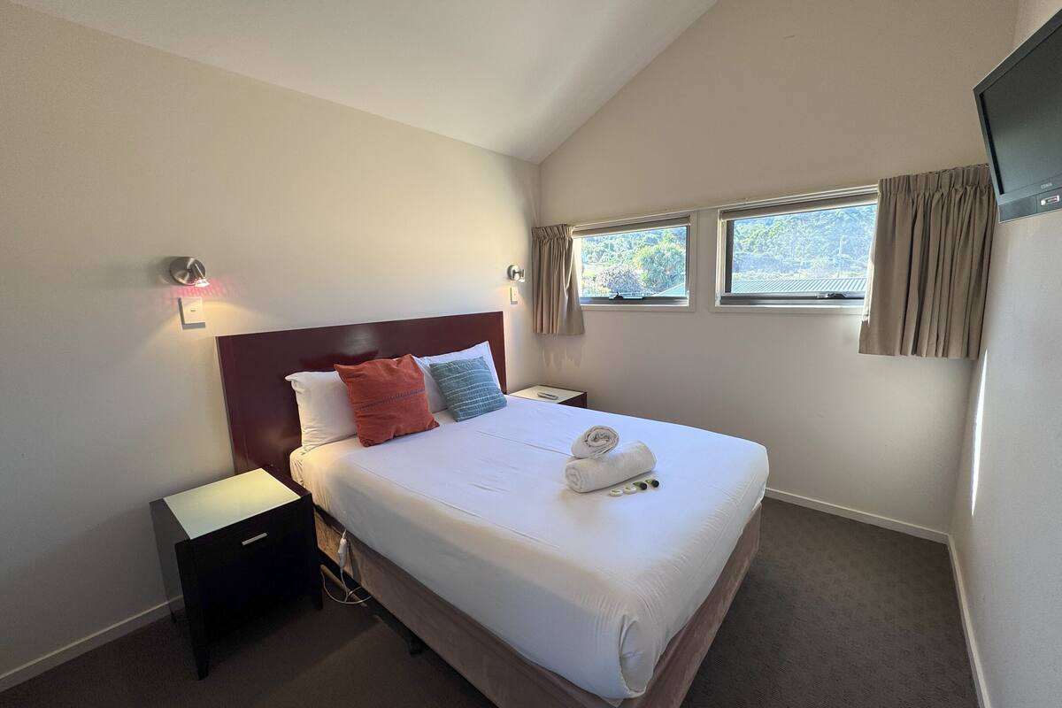 A hotel room with a bed, nightstand, and two windows with curtains.
