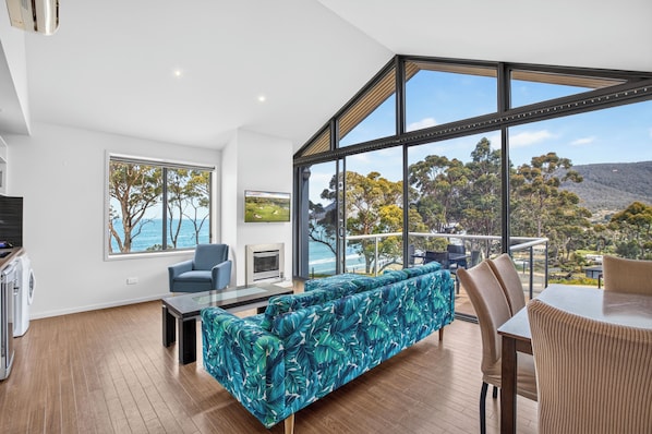 A modern living room with a large window, a sofa, a coffee table, and a dining area. - Lufra Beachside Hotel (Eaglehawk Neck)