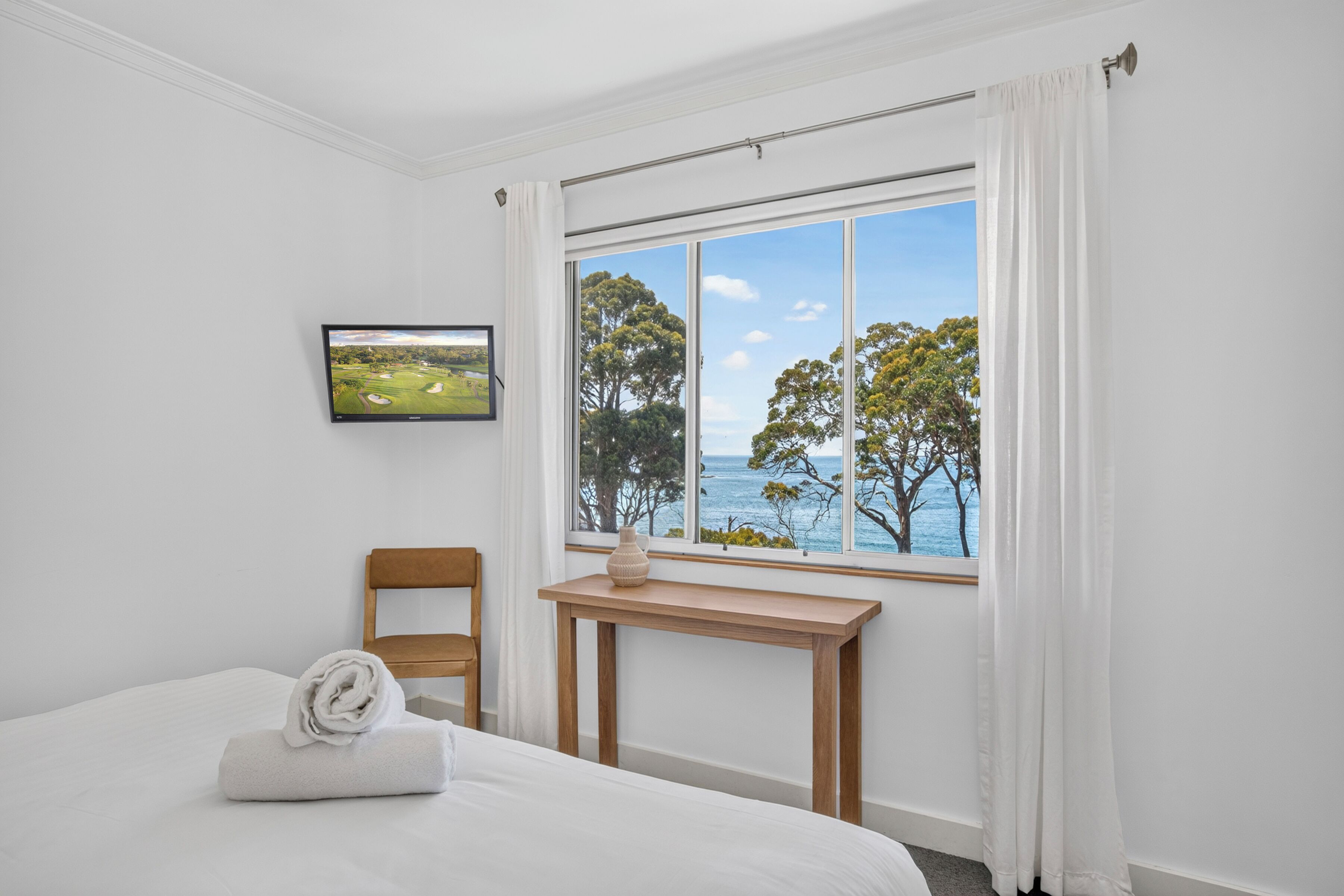 A hotel room with a bed, a wooden chair, a small table, a window with a view of trees and the sea, and a framed picture on the wall.
