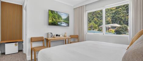 A hotel room with a bed, a desk with chairs, a television, and a window with a view of trees.