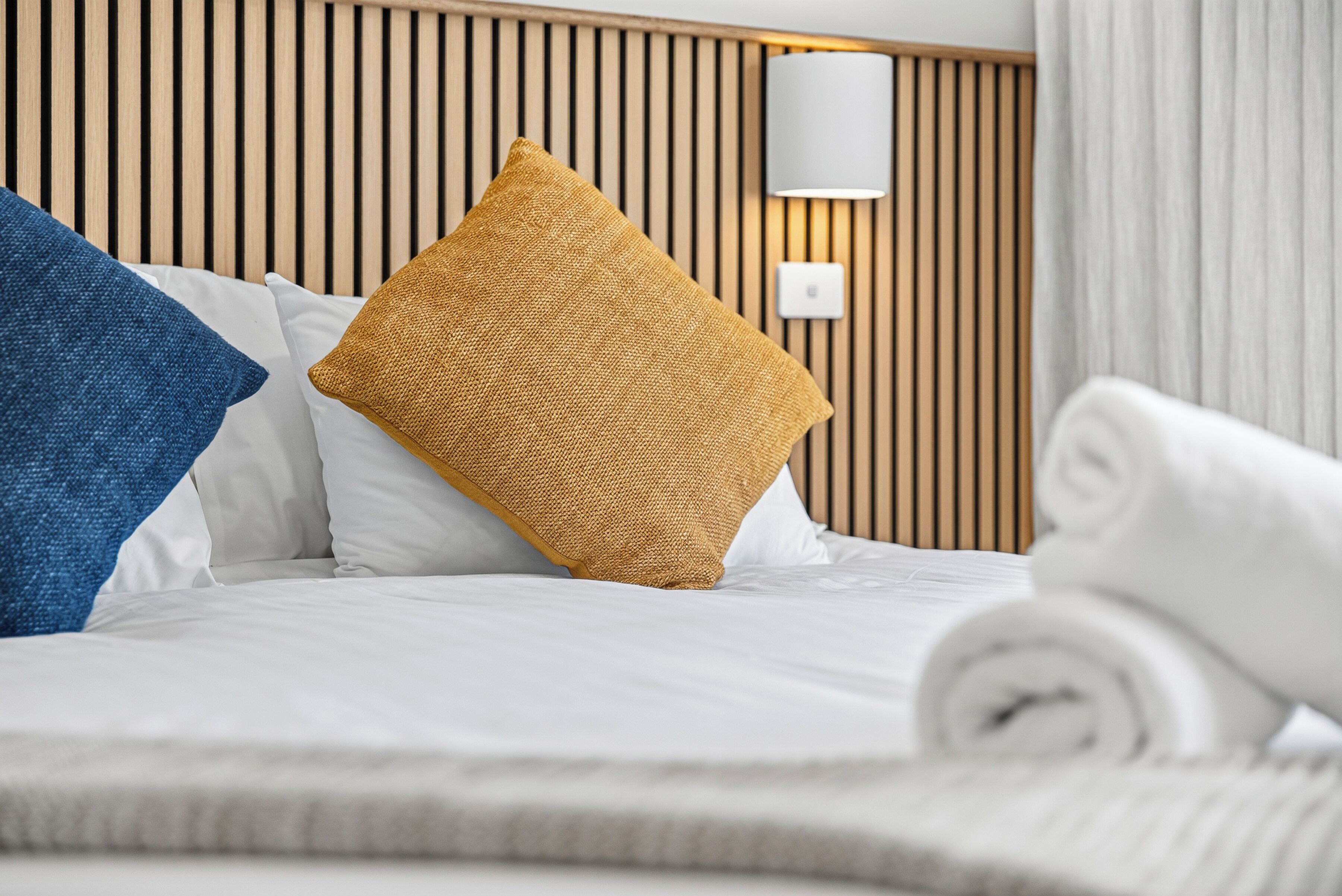 Una cama bien hecha con sábanas blancas, una almohada azul y otra mostaza, y una toalla blanca enrollada.