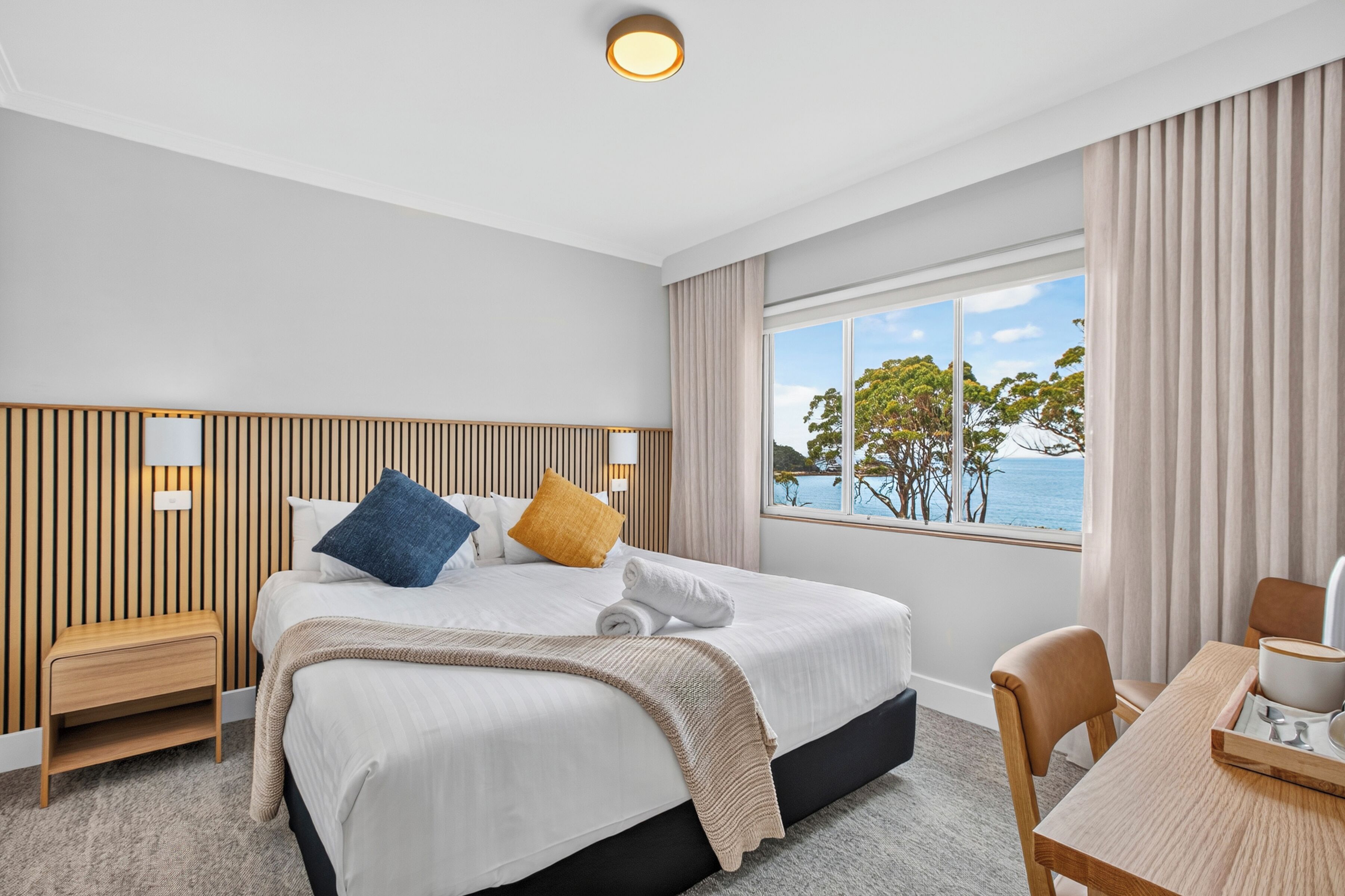 Una camera d'albergo con un letto, comodini, una scrivania con una sedia e una finestra vista mare.