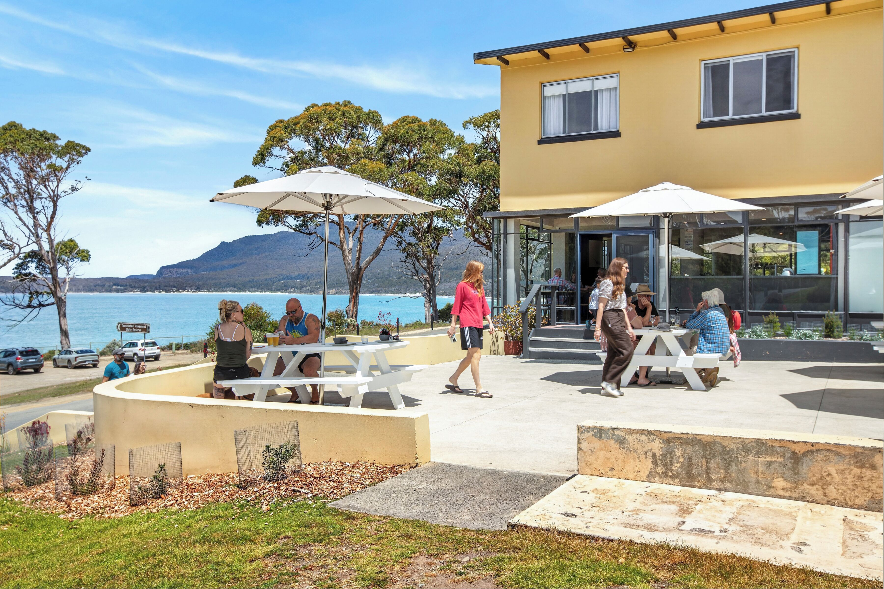 An outdoor seating area with tables and umbrellas, overlooking a body of water and hills.