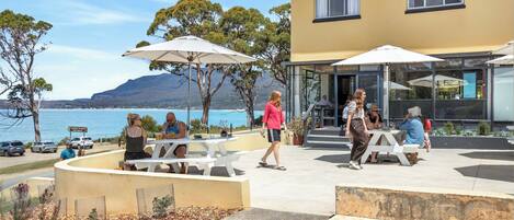 An outdoor seating area with tables and umbrellas, overlooking a body of water and hills.