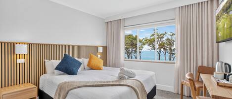 A hotel room with a large bed, a desk, a chair, and a window with a view of the sea.