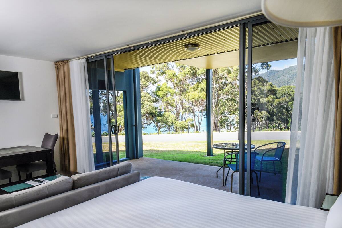 A hotel room with a bed, a desk, and a table with chairs. There is a sliding glass door leading to an outdoor area with a view of trees and hills.