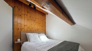 Een slaapkamer met een houten hoofdbord, een bed met witte beddengoed, een grijze plaid en een witte nachtkastje met een kopje.