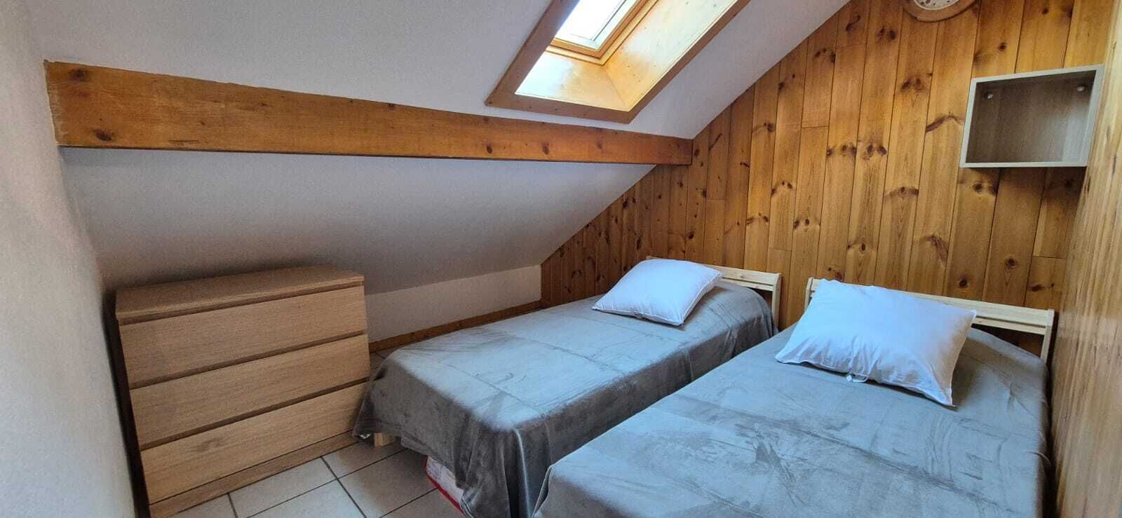 Une petite pièce avec un toit en pente, comprenant deux lits simples, une commode en bois et un puits de lumière.