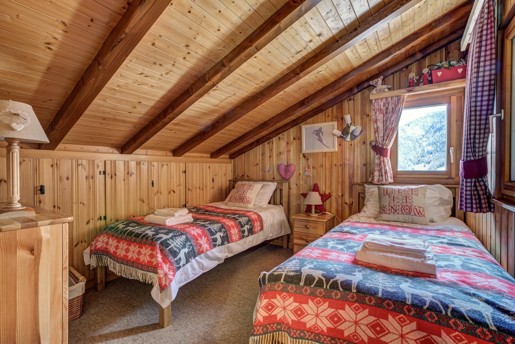 Ein gemütliches Blockhauszimmer mit zwei Betten, Holzwänden, einem Fenster mit Vorhängen und einem kleinen Tisch mit Lampe.