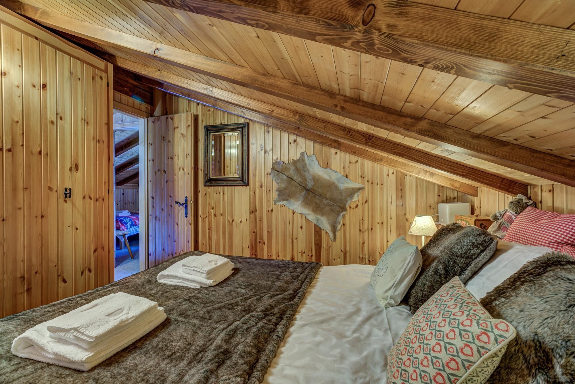 A cozy attic bedroom with wooden walls and a sloped ceiling. A bed with a blanket and pillows is visible, along with a small table and a lamp.